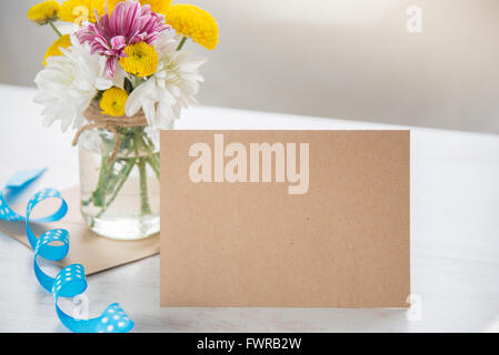 Spring flowers bouquet in a jar vase with kraft card note, envelope and blue dotted ribbon on a white wood rustic background Stock Photo