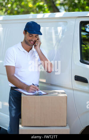 Delivery man writing in clipboard while talking on mobile phone Stock Photo