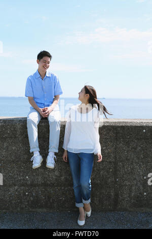 Young Japanese couple sitting on concrete wall by the sea Stock Photo