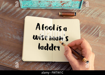 Retro effect and toned image of a woman hand writing on a notebook. Handwritten quote A fool is a wise man's ladder as inspirational concept image Stock Photo