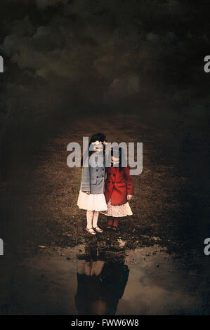 two small girls sisters standing by the puddle looking down at their reflection in water Stock Photo