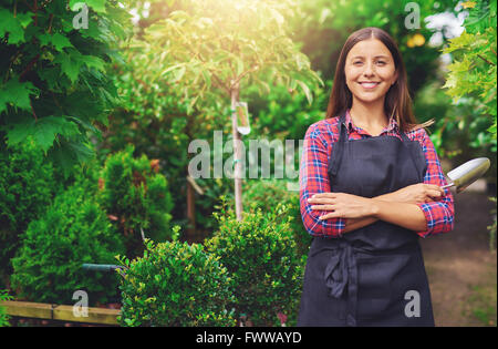 Happy attractive young female botanist working at a nursery standing in a greenhouse alongside potted topiary trees with a trowe Stock Photo