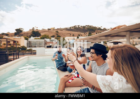 Outdoor shot of happy young people sitting on the edge of the pool having a celebratory toast. Friends raising a toast during a Stock Photo