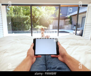 POV shot of man using digital tablet while lying on bed. Human hand holding digital tablet with blank screen. Stock Photo