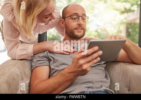 Mature man sitting on sofa using digital tablet with his girlfriend standing by with her hands on his shoulders. Couple at home Stock Photo