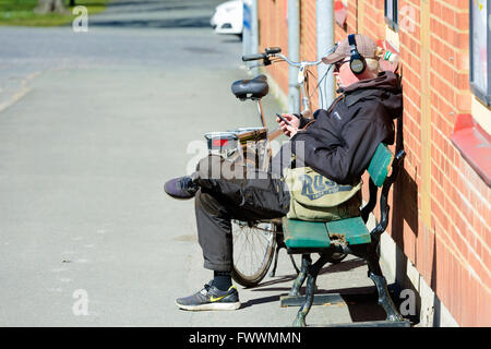 Simrishamn, Sweden - April 1, 2016: Man sitting on a bench next to a wall listening to something through earphones. Bike is park Stock Photo