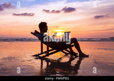 silhouette of successful business man reading emails on laptop on the beach at sunset, freelance job concept, work abroad Stock Photo