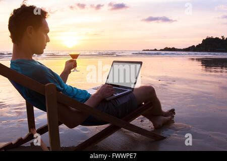 young business man with cocktail working on laptop on the beach at sunset, freelance job online, focus on the screen Stock Photo