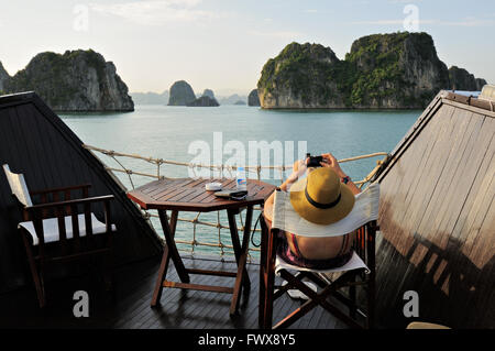 Young caucasian woman sitting on a chair takes a picture in Bai Tu Long area of Ha Long Bay from the deck of a junk, Vietnam Stock Photo