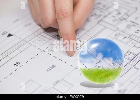 Finger from an unknown person is showing at the plan of the house. Blue sky, blueprint and green grass are in the glass ball. Stock Photo