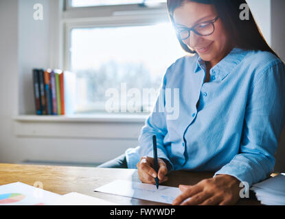 Selective focus close up on happy entrepreneur female wearing glasses and blue shirt at desk writing on form next to cup of coff Stock Photo