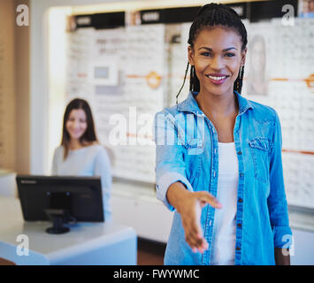 Pretty young African American optometrist greeting a customer at the door with a beaming smile and outstretched hand or offering Stock Photo