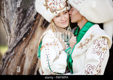 Couple in ethnic costumes embrace on background of textured wood, groom kisses bride at cheek. Stock Photo