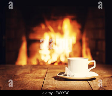 White cup of tea with teabag on wooden table near  fireplace. Winter and Christmas holiday concept. Photo with retro filter effe Stock Photo