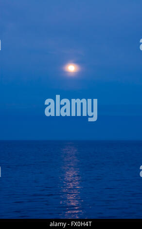 Soft moon in clouds and haze reflected in water on blue evening sky. Stock Photo