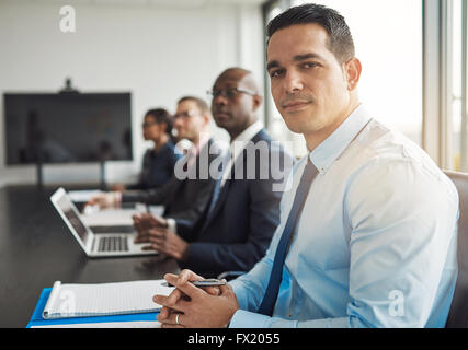 Young Hispanic businessman in a meeting with co-workers at the office sitting at a conference table turning to smile at the came Stock Photo