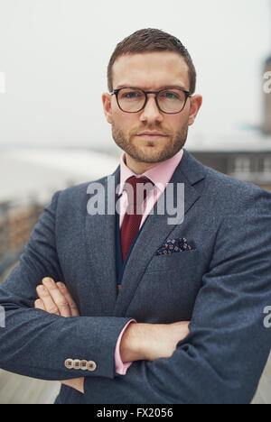 Serious determined young business man wearing glasses standing with folded arms looking at the camera with a pensive intense exp Stock Photo