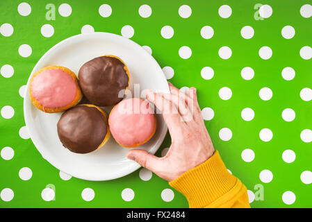 Male hand picking donut with sweet strawberry topping from a plate, top view of homemade tasty donuts on kitchen table polka dot Stock Photo