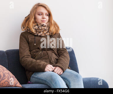 Blond Caucasian teenage girl in warm clothes sitting on sofa over white wall Stock Photo