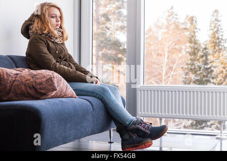 Beautiful blond Caucasian teenage girl in warm clothes sitting on sofa near the window in hallway Stock Photo