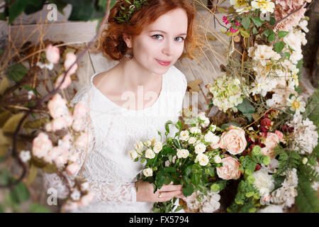 Young red-haired bride in a green wreath is holding a bouquet of white roses and gently smiles. She is surrounded by flowers and Stock Photo