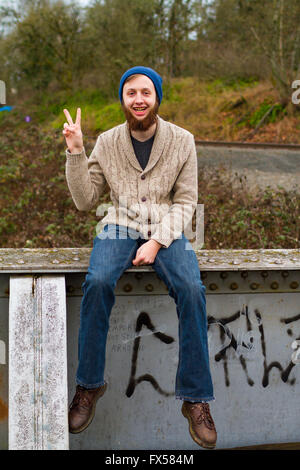 Hippie man sitting on a bridge giving a peace sign in this trendy hipster fashion portrait. Stock Photo