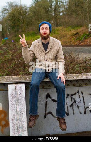 Hippie man sitting on a bridge giving a peace sign in this trendy hipster fashion portrait. Stock Photo