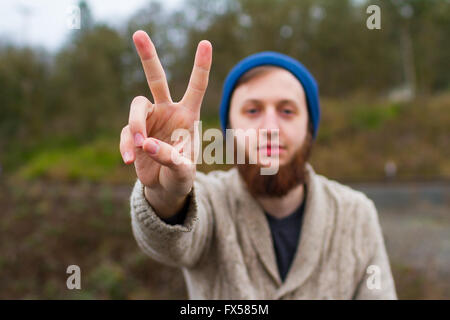 Hippie man sitting on a bridge giving a peace sign in this trendy hipster fashion portrait. Stock Photo