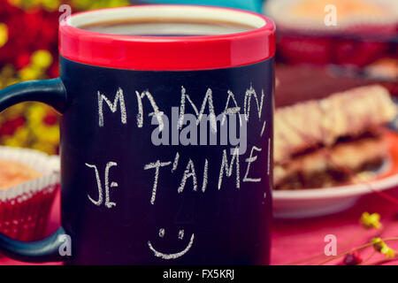 the sentence maman je t aime, I love you mom in french handwritten with chalk in a black mug with coffee, with some muffins in t Stock Photo