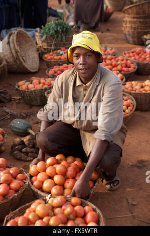 Portrait of a young Ugandan vendor wearing a yellow bucket hat crouching behind baskets of fresh tomatoes at an outdoor market in Uganda Stock Photo