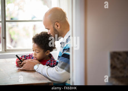 Mixed race father and son using cell phone at table Stock Photo