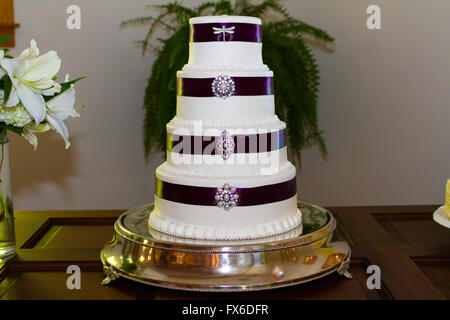 Tall white wedding cake wrapped in purple ribbon with brooches attached to each tier of the dessert. Stock Photo