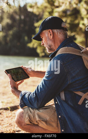 Close up portrait of senior man with backpack sitting by a lake and using digital tab in the forest for navigation. Stock Photo