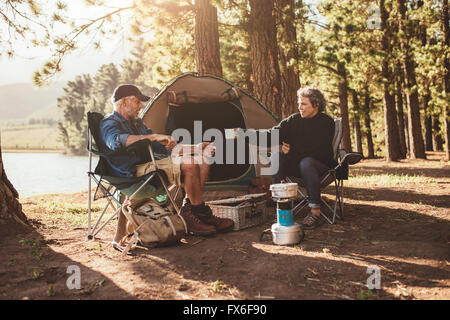 Portrait of senior couple camping by a lake, with woman giving a cup of coffee to man. Stock Photo