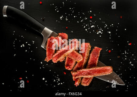 Medium rare steak slices on a knife on black plate, with salt flakes and peppercorns Stock Photo