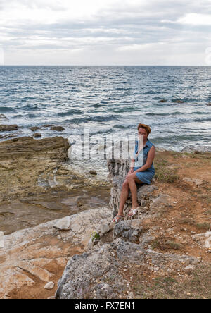 Middle aged caucasian tanned woman in jeans dress sitting on the rocky sea beach. Istria, Croatia. Stock Photo