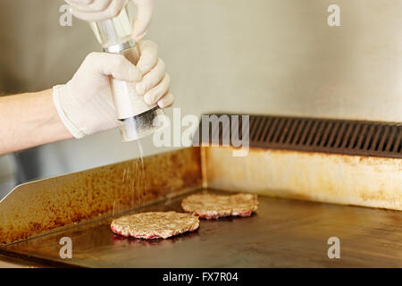 Gloved hands grinding salt onto two frying hamburger patties Stock Photo