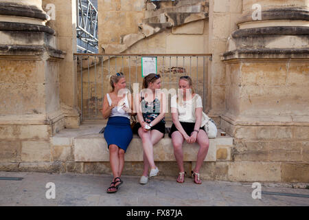 Three tourists take a break from touring in Valletta by sitting outside the ruins of the Royal Opera House. Stock Photo
