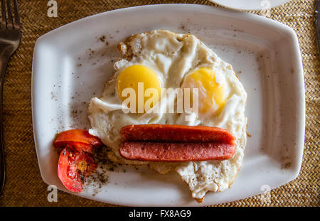 Smiley face breakfast of sunny side eggs and a split sausage with tomato and black pepper garnish on a white plate