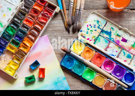 Set of watercolor paints, brushes for painting and paper sheet of painting on old wooden background. Top view. Stock Photo