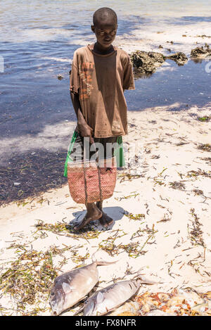 Pemba, Zanzibar, Tanzania - 10 October 2015: Young African boy with a colourful bag standing in front of freshly caught fish on Stock Photo