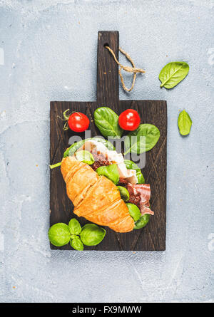 Croissant sandwich with smoked meat Prosciutto di Parma, sun dried tomatoes, fresh spinach and basil on dark wooden board over s Stock Photo