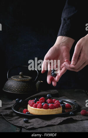 Female hands decorating by berries tart with custard, raspberries and blueberries, served on vintage tray with iron teapot on te Stock Photo
