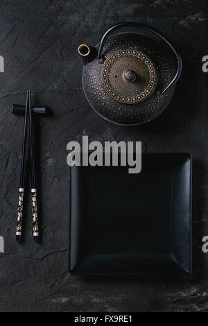 Black painted chopstics on chopsticks rest, empty square plate and iron teapot over black textured surface. Flat lay. Stock Photo