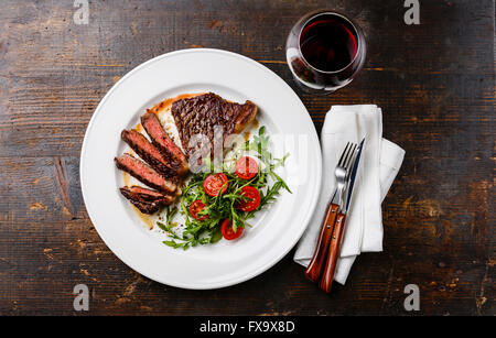 Sliced medium rare grilled Beef steak Striploin, salad with tomatoes and arugula on white plate and red wine on dark wooden back Stock Photo