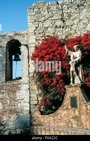 Bright bougainvillea flowers surround a statue of Spanish missionary Junipero Serra embracing a young Indian boy, a 1914 monument by American artist Tole Van Rensaalar in the courtyard of Mission San Juan Capistrano in Orange County, California, USA. The fabled Franciscan friar founded the mission in 1776, the seventh of 21 California missions established up and down the state to expand Spain’s territory and convert the Native Americans to Christianity. Father Serra was canonized as a Catholic saint by Pope Francis in 2015. Stock Photo