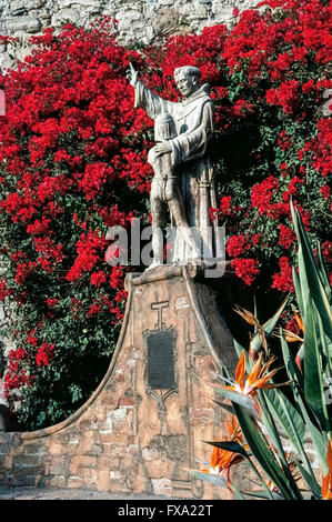 Bright bougainvillea flowers surround a statue of Spanish missionary Junipero Serra embracing a young Indian boy, a 1914 monument by American artist Tole Van Rensaalar in the courtyard of Mission San Juan Capistrano in Orange County, California, USA. The fabled Franciscan friar founded the mission in 1776, the seventh of 21 California missions established up and down the state to expand Spain’s territory and convert the Native Americans to Christianity. Father Serra was canonized as a Catholic saint by Pope Francis in 2015. Stock Photo