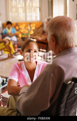 Old people in geriatric hospice: young attractive hispanic woman working as nurse takes care of a senior man on wheelchair. She Stock Photo