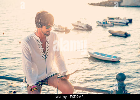 young man listening music with headphones from tablet in a vacations destination, sitting by a little port Stock Photo