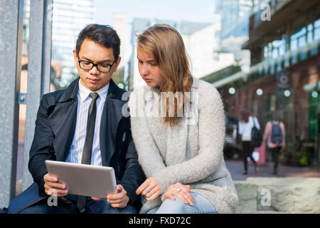 Two serious young people looking into digital tablet sitting outside on city street Stock Photo